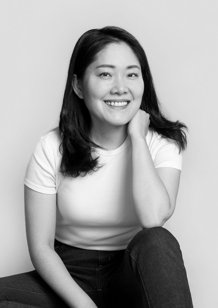Black and white portrait of a woman with shoulder-length dark hair, wearing a white top. She has a calm and confident expression, looking directly at the camera. The background features a textured brick wall.